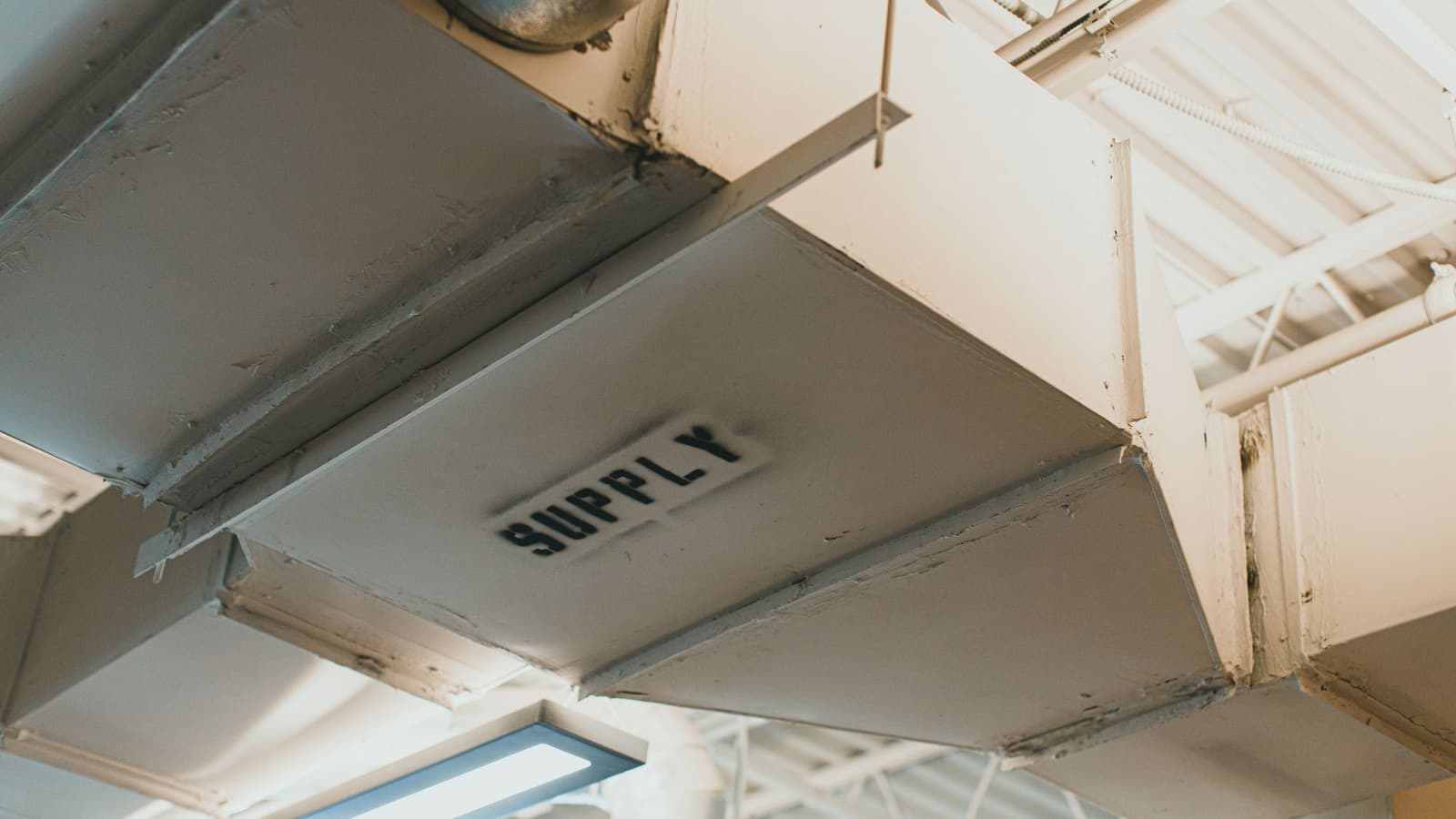 Painted sheet-metal rectangular supply duct running along a bright ceiling